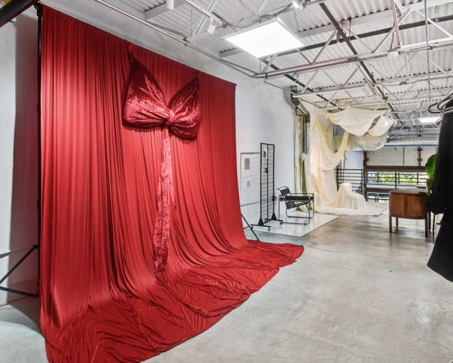 The image shows a spacious studio with a large red fabric backdrop styled as a bow, complemented by draped white fabric in the background and various furniture pieces arranged throughout the space.