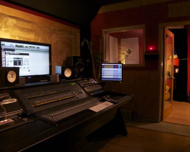 The image shows a professional music recording studio with a control console, computer monitors, and a hallway leading to a room with guitars.