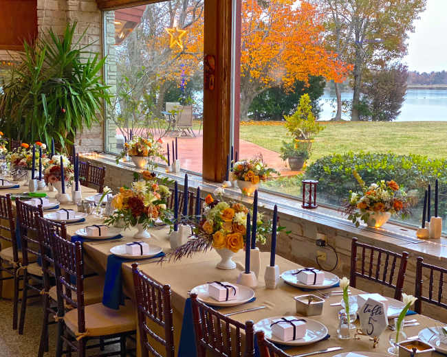 A long dining table is set with floral centerpieces and candles, overlooking a lake with autumn trees in the background.