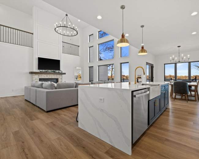 A modern kitchen features an island with a white marble countertop, pendant lights, and a spacious living area with large windows overlooking a scenic view.