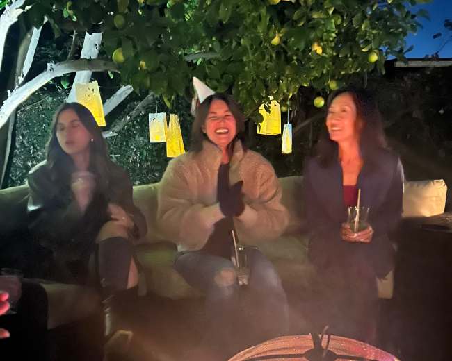 Three women sit on a couch around a fire pit, smiling and engaged in conversation under a tree illuminated by hanging lanterns.