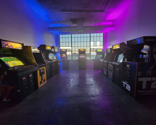 A room filled with several retro arcade machines arranged symmetrically around a central area, illuminated by purple and blue lights.