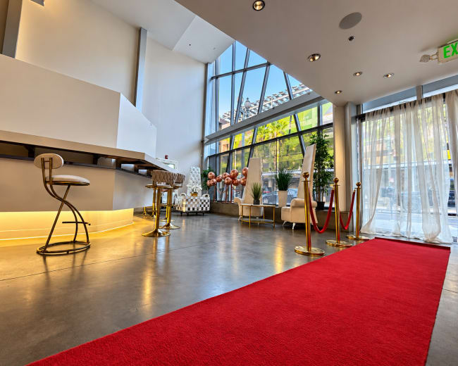 The image shows a modern lobby featuring a red carpet, bar stools, plants, and large windows letting in natural light.