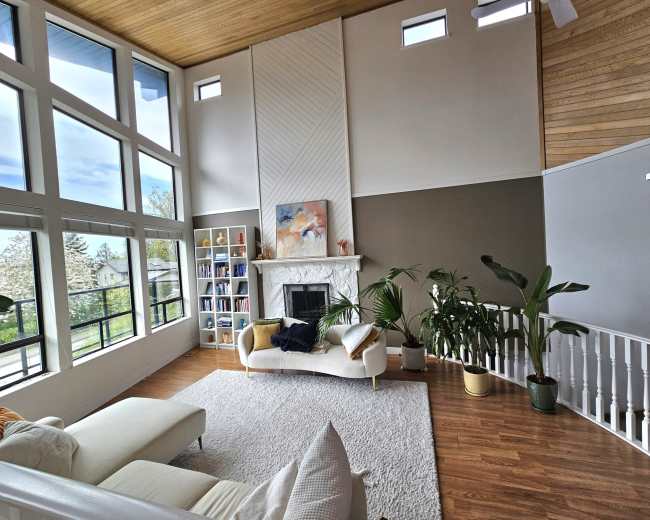 A bright living room features large windows, a fireplace, a bookshelf, and a sofa with plants and a rug on the wooden floor.