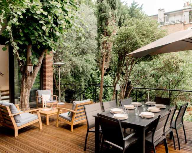 A wooden deck features a large dining table surrounded by chairs, with a shaded seating area and greenery visible in the background.
