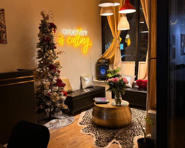The image shows a cozy interior space with a decorative Christmas tree, a neon sign that reads "Creativity is calling," and a round table adorned with flowers.