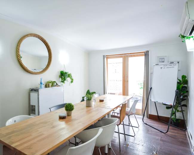 A bright meeting room features a wooden table surrounded by chairs, a mirror on the wall, and a whiteboard near a window.