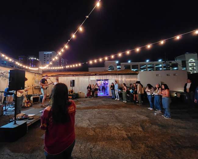 A musician performs on a rooftop stage in front of a small crowd under string lights at night.