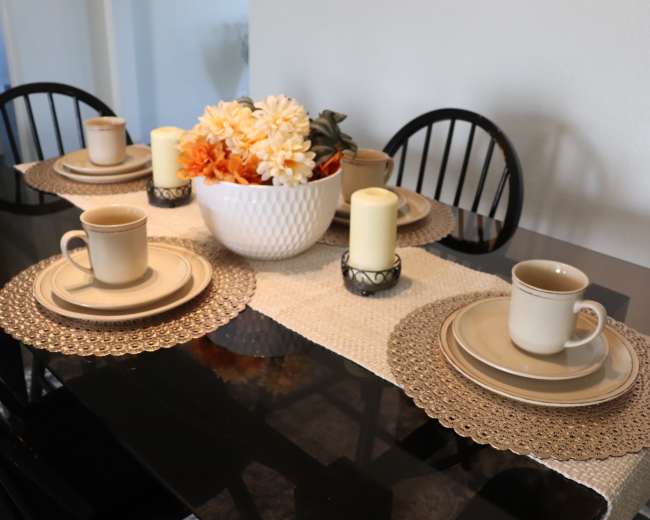 A dining table is set with beige plates and mugs, adorned with a floral centerpiece and lit candles on a runner.