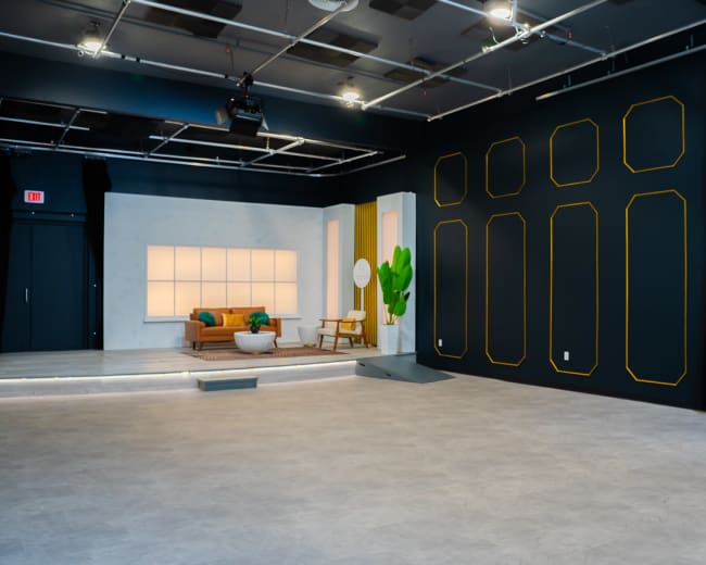 The image shows a modern studio space with a light-colored seating area against a backdrop of dark walls featuring geometric outlines and a wall with black curtains.