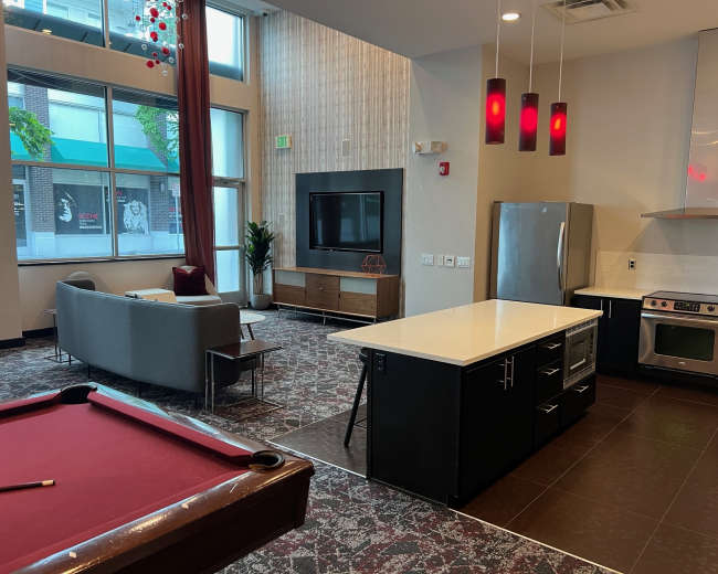 The image shows a modern lounge area featuring a gray couch, a pool table, a kitchen island, and large windows that illuminate the space.