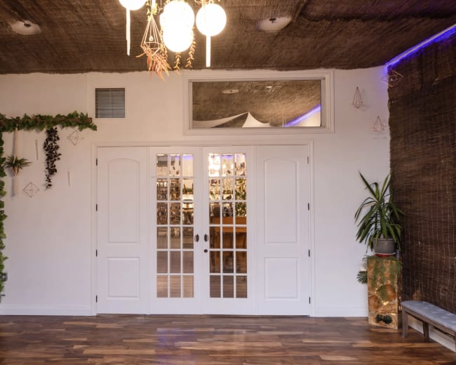 The image shows a bright indoor space featuring double doors, a wooden floor, and decor elements like plants and a woven ceiling.