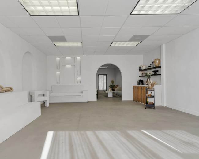 The image shows a minimalistic, white interior space featuring a raised platform, an archway, and simple furniture arrangements.