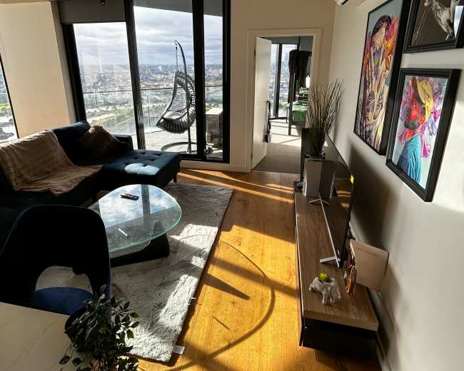 The image depicts a modern living room with a sectional couch, a glass coffee table, a flat-screen TV on a wooden stand, and large windows showcasing a view of the city.