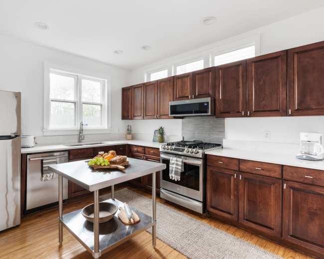 A kitchen with wooden cabinets, a stainless steel refrigerator, and a central island with fruits and a bowl, illuminated by natural light from the windows.