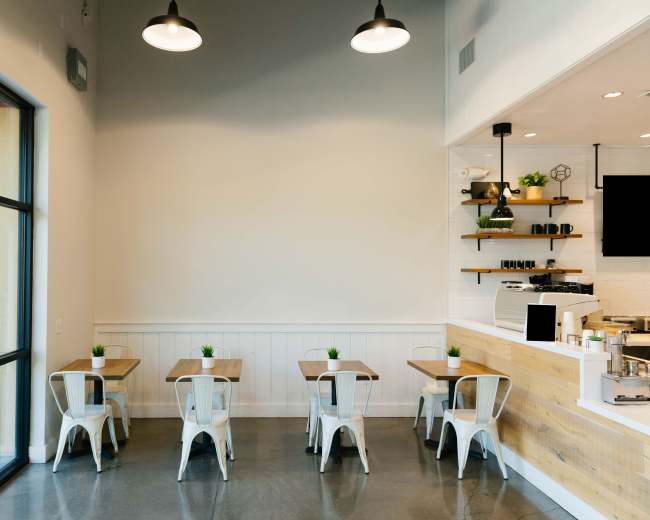 The image shows a minimalistic cafe interior with four wooden tables, metal chairs, and a counter area featuring shelves and a coffee machine.