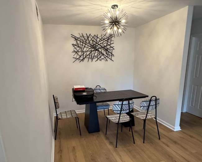 A dining area features a black table surrounded by three striped chairs, with a decorative wall piece and a modern light fixture above.