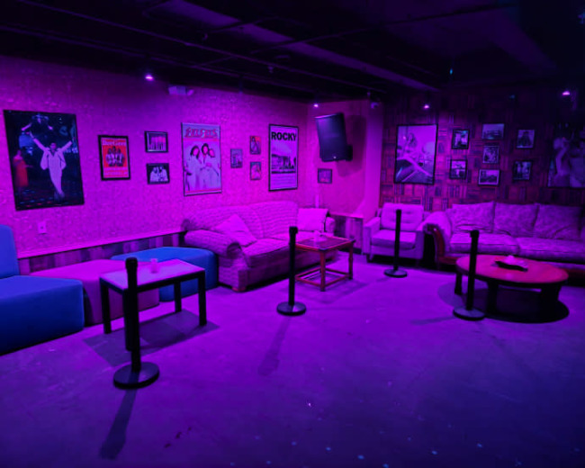 The image shows a dimly lit lounge area with purple lighting, featuring couches, tables, and framed posters on the walls.