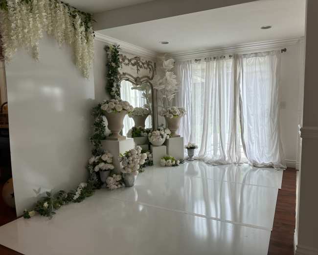 A bright room features white walls and glossy floors, adorned with floral arrangements and decorative mirrors, alongside sheer curtains.