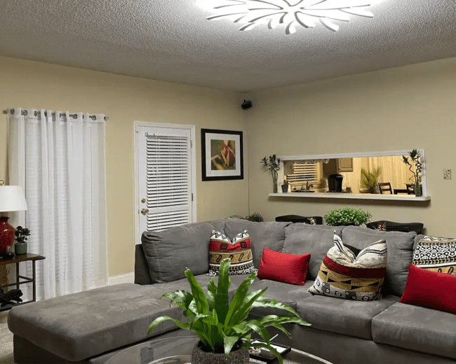 A cozy living room with a gray sectional sofa adorned with colorful pillows, a round glass coffee table, and a wall-mounted light fixture above.