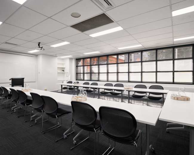 The image shows a modern conference room with several white tables arranged in a U-shape and black chairs, illuminated by natural light from large windows.