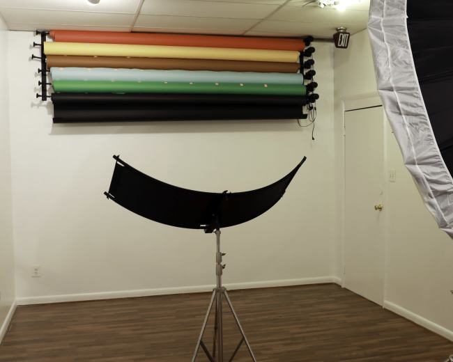 A photography studio with backdrop rolls hanging on the wall and a large umbrella softbox on a stand.