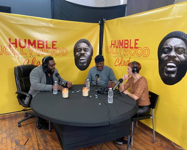 Three people sit at a table during a podcast recording, with yellow walls featuring black-and-white portraits in the background.