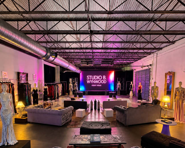 The image shows a spacious studio with a polished concrete floor, stylish seating areas, and various fashion garments displayed on racks and mannequins, illuminated by vibrant lighting.
