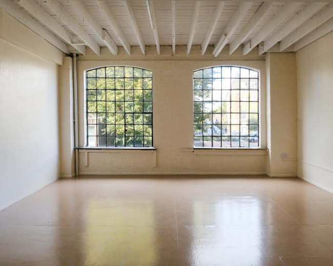 The image shows a bare room with two large arched windows and polished floors, illuminated by natural light.