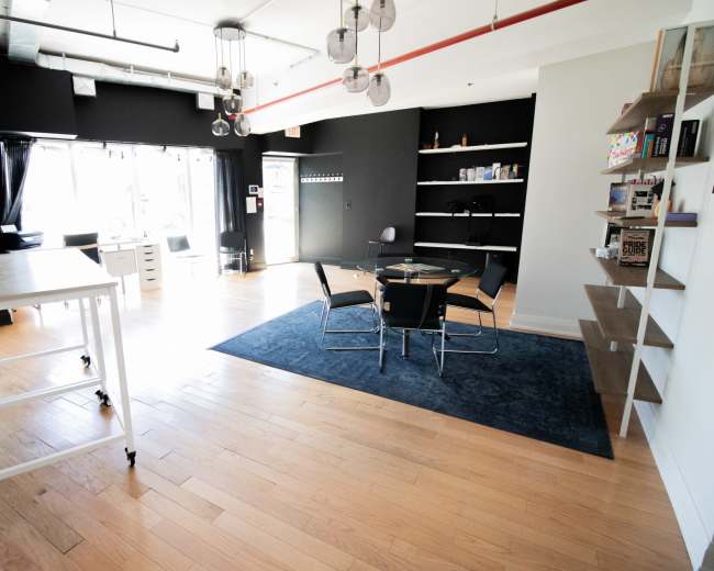 The image shows a modern office space featuring a circular glass table surrounded by chairs, hardwood flooring, and large windows allowing natural light.