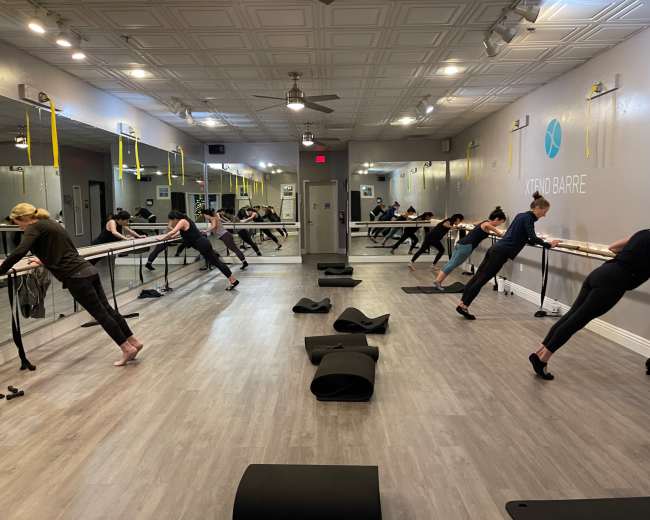 A group of individuals participates in a barre fitness class in a mirrored studio with exercise mats on the floor.