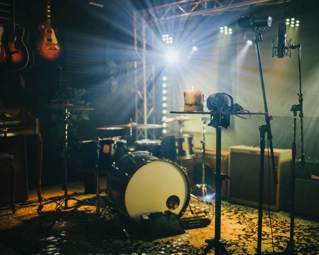 The image shows a music studio setup with a drum kit, microphones, a drink on a stand, and various instruments illuminated by stage lights.