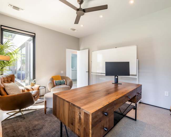 A modern home office featuring a large wooden desk, a comfortable seating area with a chair, a wall-mounted whiteboard, and a guitar leaning against the wall.