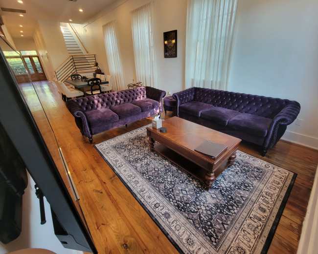 The image shows a living room with two purple velvet sofas, a wooden coffee table, and a patterned area rug on polished wooden flooring, leading to a staircase in the background.