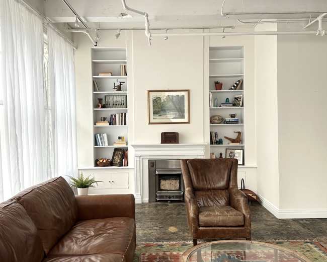 The image shows a modern living space with a brown leather sofa and a matching armchair, alongside white bookshelves and a fireplace.