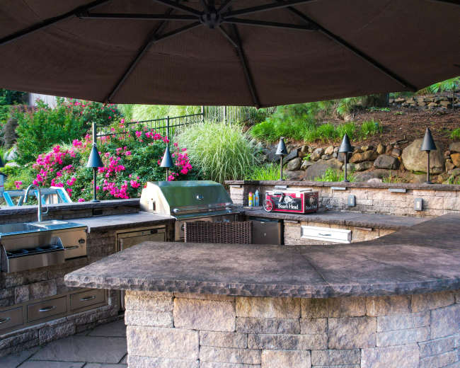 The image shows an outdoor kitchen setup with a stone countertop, grill, and shaded seating area surrounded by lush greenery and flowering plants.