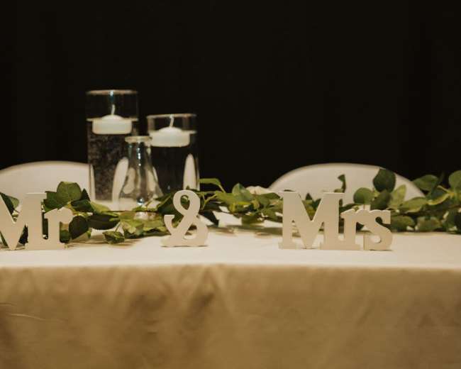 A decorated table with the words "Mr" and "Mrs" placed between two glass vases containing water and floating candles, all surrounded by a green garland.
