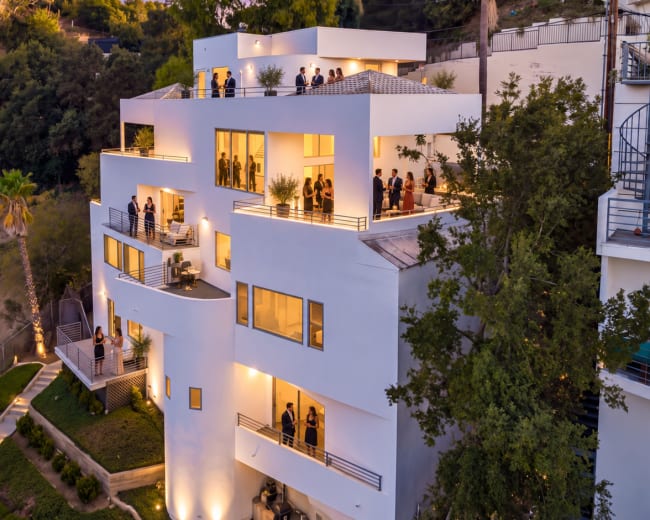 A modern three-story building with large windows and balconies is surrounded by greenery and features people socializing on its terraces during twilight.