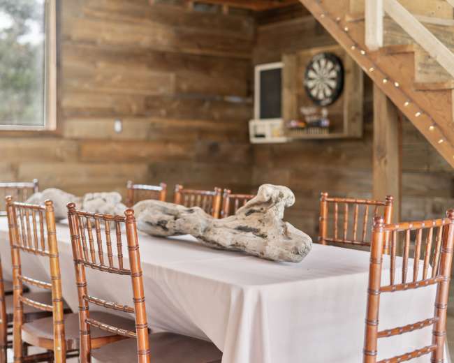 A long table with a white tablecloth is set in a wooden room, surrounded by multiple wooden chairs, and features a decorative piece of driftwood at the center.
