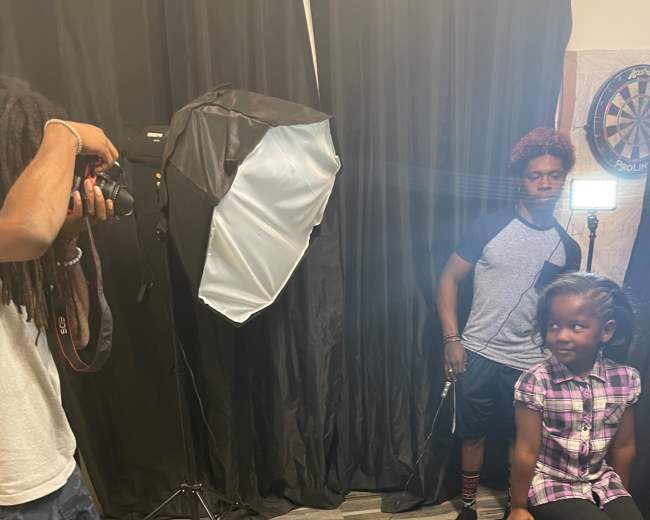 A photographer captures an image of a young girl sitting on a stool in a studio setting with professional lighting.