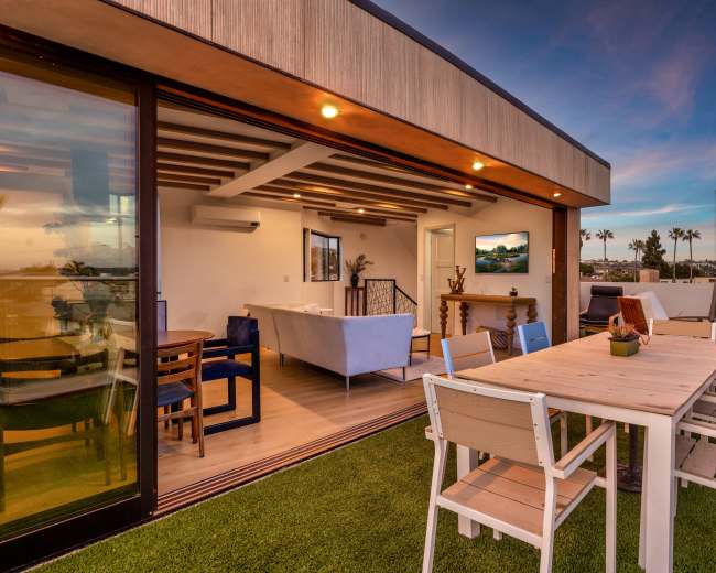 A modern rooftop terrace features a dining table surrounded by white chairs, overlooking a sunset with palm trees in the distance.