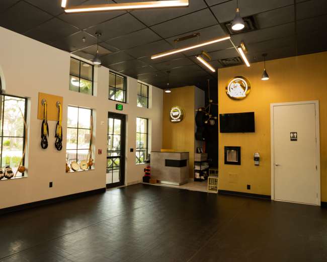 The image shows a fitness studio interior with large windows, black flooring, and exercise equipment such as TRX straps and kettlebells displayed on the walls.