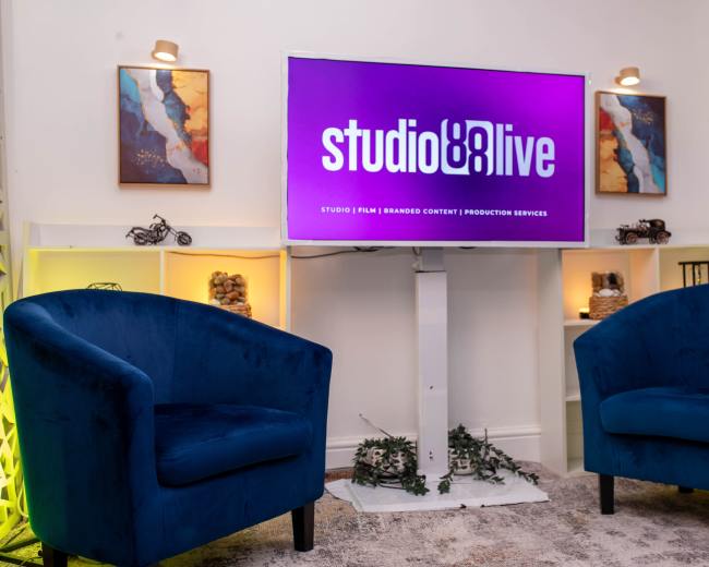 The image shows a modern setup featuring a television displaying the logo "studio & live" above two blue velvet chairs, with decorative shelves on either side.