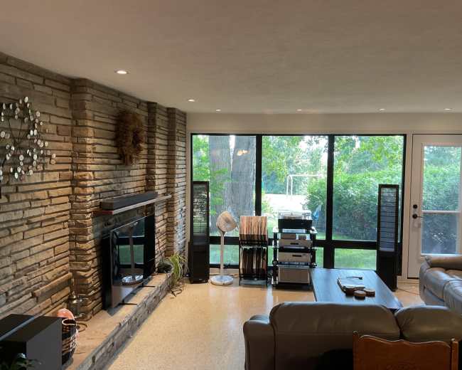 The image shows a cozy living room with a stone fireplace, a seating area, and large windows overlooking a green garden.