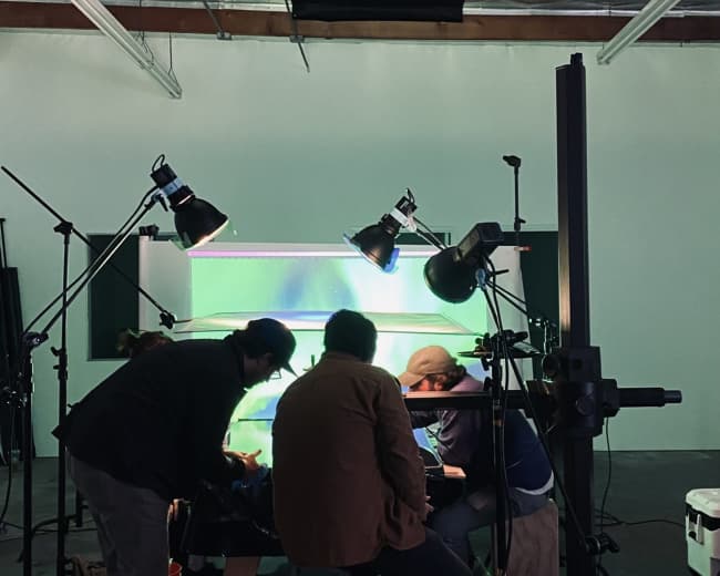 A group of people is gathered around a table in a studio, working on a project illuminated by multiple overhead lights.