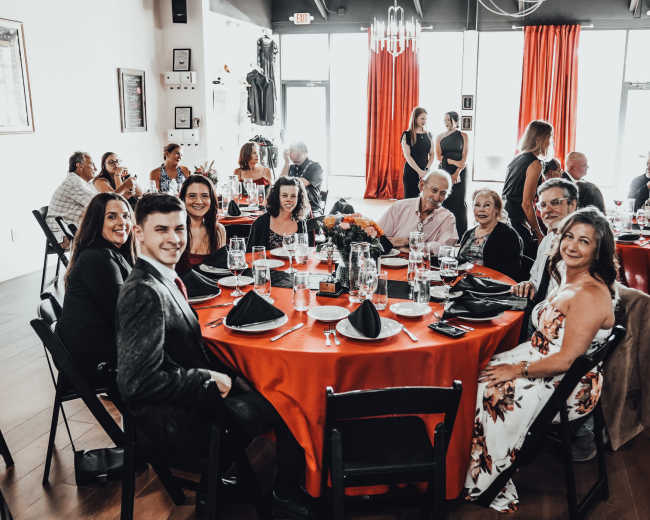 A group of people is seated around a round table dressed in a red tablecloth at a social gathering in a well-lit venue.
