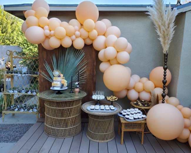 The image shows a decorated outdoor space featuring a large arrangement of peach-colored balloons, two woven tables with a tiered cake and assorted desserts.