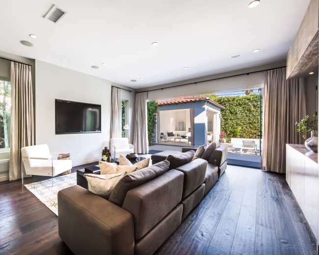 The image shows a modern living room with a dark leather sofa, a large television mounted on the wall, and sliding glass doors leading to an outdoor area.