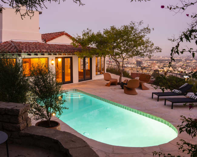 The image shows a clear swimming pool surrounded by a stone patio and lounge chairs, with a house nearby and a city skyline visible in the distance at twilight.