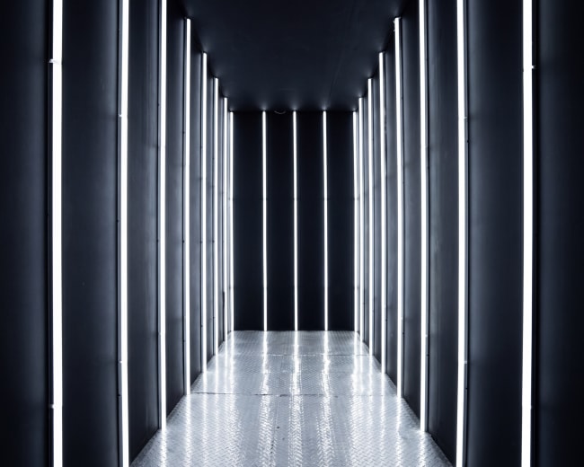 The image shows a narrow, dark corridor lined with vertical fluorescent lights and a metallic patterned floor.